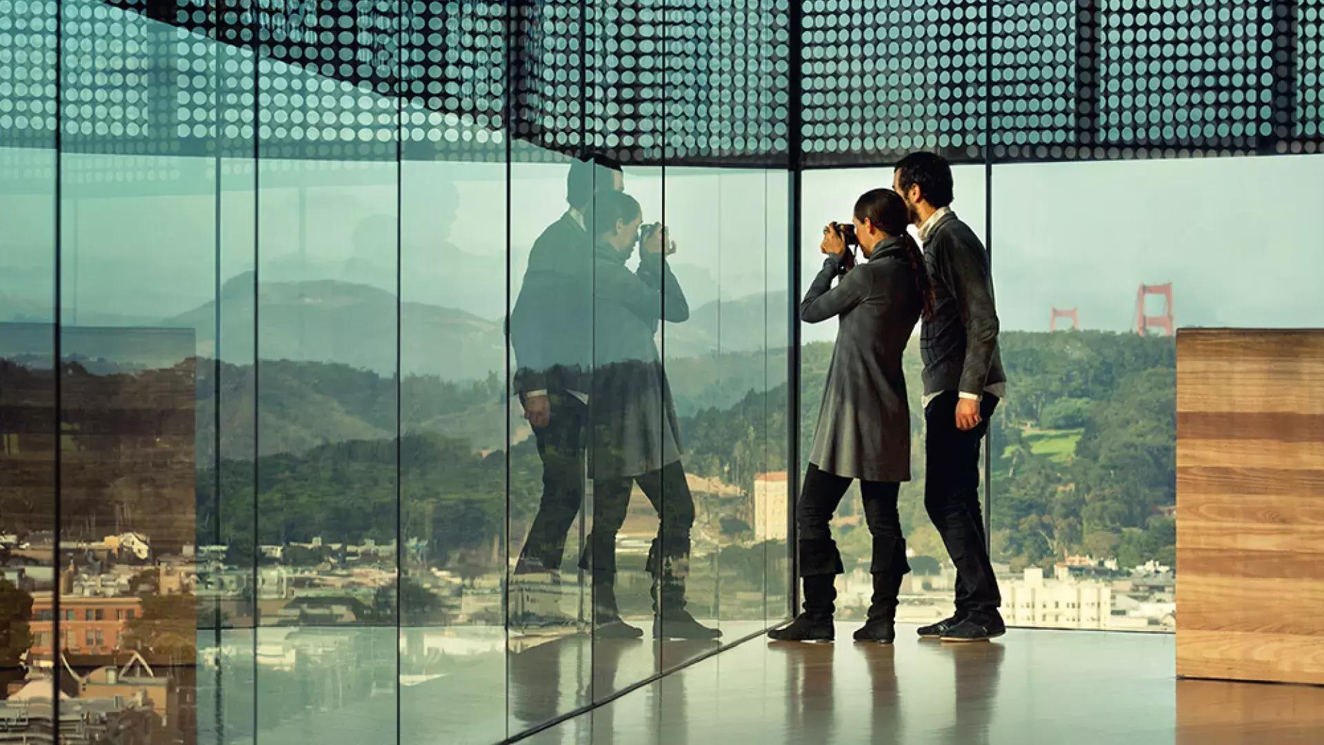 Two people peer through glass walls at the de Young Museum in San Francisco.