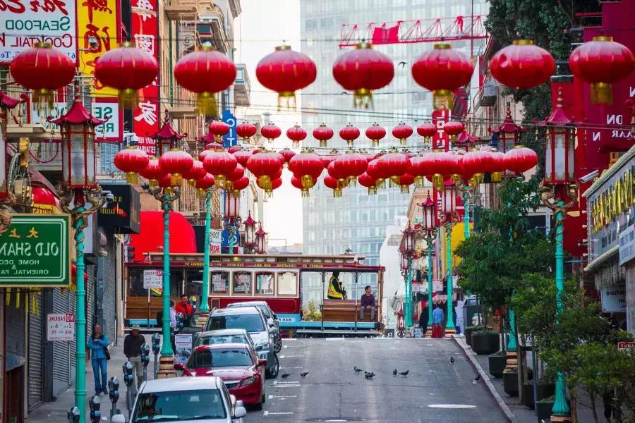 Une rue vallonnée du quartier chinois de San Francisco est représentée avec des lanternes rouges suspendues et un tramway qui passe.