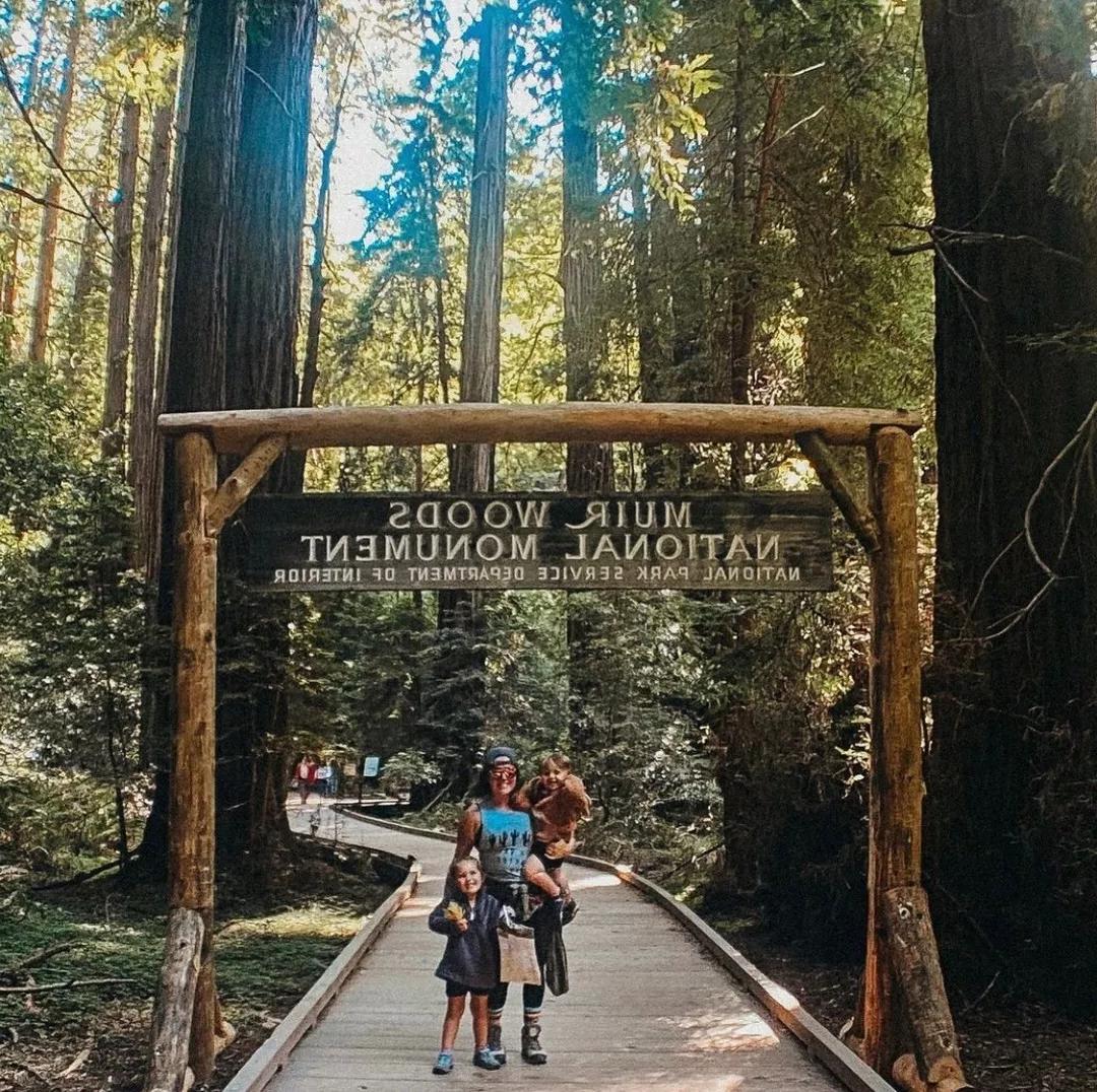 Woman and children stand in front of Muir Woods National Monument signage on a wooden trail in a forest