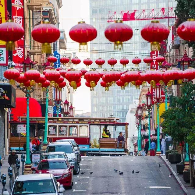 A hilly street in San Francisco's Chinatown is pictured with red lanterns dangling and a streetcar passing by.